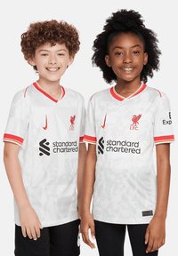 Maglietta bianca del Liverpool FC con accenti rossi, caratterizzata da un colletto a V, logo, testo dello sponsor e un motivo testurizzato. Indossata da un ragazzo e una ragazza.