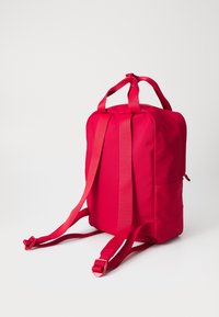 Sac à dos rouge avec deux poignées supérieures et des bretelles réglables. Il possède une texture lisse et une poche zippée sur un côté.