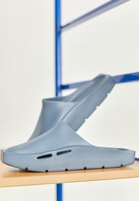 Zoccoli blu senza lacci realizzati in materiale gommoso testurizzato, dotati di fori di ventilazione e un design minimalista con punta tonda e suola piatta.