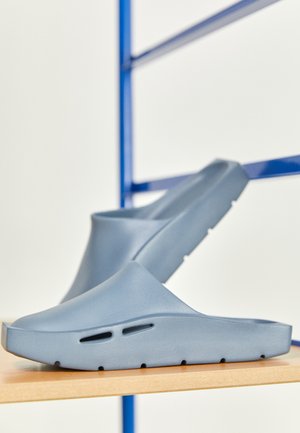 Zuecos azules de tela de goma texturizada, con agujeros de ventilación y un diseño minimalista con punta redondeada y suela plana.