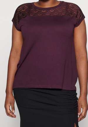 Donna che indossa una maglia prugna a maniche corte con dettaglio in pizzo sullo scollo e una gonna nera arricciata con una spacco laterale.