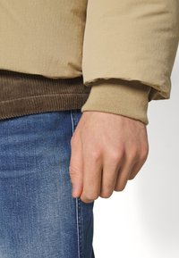 Beige padded jacket with a textured finish and ribbed brown hem, layered over a brown corduroy top. Hand clenched on blue denim.