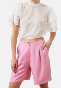 Blusa trasparente bianca a pois con maniche a volant, abbinata a pantaloni corti a vita alta rosa plissettati con chiusura a bottone e tasche laterali.