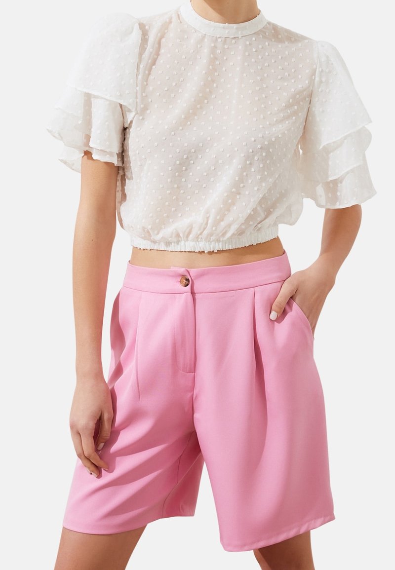 Blusa trasparente bianca a pois con maniche a volant, abbinata a pantaloni corti a vita alta rosa plissettati con chiusura a bottone e tasche laterali.