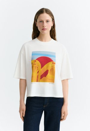 Femme aux cheveux longs porte un t-shirt blanc oversize avec un motif de chaises jaunes et un cercle rouge sur fond bleu, associé à un jean foncé.