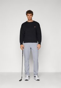 Svart strikket genser med ribbestrikket nederkant, grå bukser og hvite treningssko. Modellen holder en golfkølle med et mønstret grep.