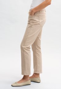Pantaloni in cotone beige con vestibilità dritta, tasche laterali e cuciture in vista. Indossati con scarpe basse di colore chiaro.