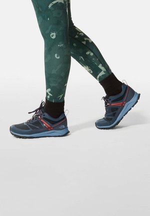Nogi w ciemnozielonych legginsach z wzorem, czarne skarpetki i niebiesko-czerwone buty trekkingowe North Face stawiają kroki do przodu na jednolitym białym tle.