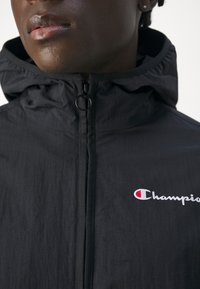 Giacca nera con cappuccio e zip frontale, presenta un logo Champion in bianco e rosso. Il tessuto ha una texture liscia e leggera.