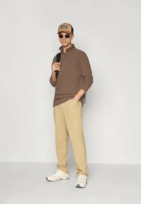 Καφέ πουλόβερ με μακριά μανίκια, μπεζ παντελόνι, καπέλο σε απόχρωση ταν με λογότυπο, λευκά παπούτσια και γυαλιά ηλίου. Το σύνολο έχει χαλαρό σχέδιο.