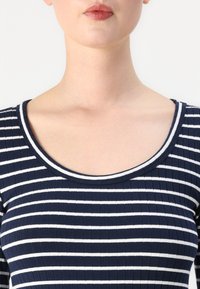 Ναυτικό μπλε ριγωτό τοπ με κάθετες λευκές λωρίδες, διακοσμημένο με φαρδιά γύρο λαιμό και λεία υφή. Απεικονίζεται η περιοχή του άνω στήθους.