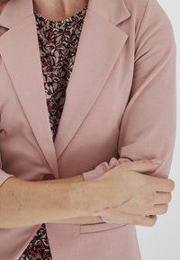Světle růžový blazer se strukturovaným designem, vybavený šikmým klopem a květinovým vzorem pod ním. Ruka vykazuje jemné jizvy.