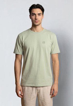 Junger Mann mit kurzen dunklen Haaren, der ein schlichtes hellgrünes T-Shirt und beige Hosen trägt und vor einem neutralen Hintergrund steht.