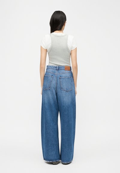 Γυναίκα με μακριά σκούρα μαλλιά, φορώντας ένα λευκό κοντομάνικο πουκάμισο κάτω από γκρι αμάνικο τοπ και μπλε τζιν παντελόνι με φαρδιά μπατζάκια, στέκεται με την πλάτη προς τα εμπρός.