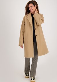 Vrouw in een beige lange jas, zwart topje, met een patroon op haar broek en zwart-witte sneakers, die glimlacht en haar haar aanraakt.