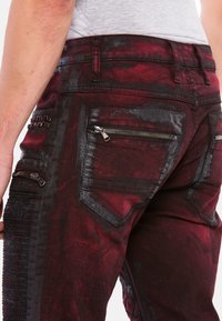 Rote und schwarze Denim-Jeans mit mehreren Reißverschlusstaschen, strukturierten Akzenten und einem distanzierten, malerischen Muster auf den Beinen.