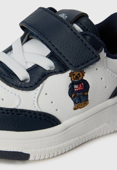 Zapatilla con upper de cuero blanco y acentos en azul marino, material texturizado, tira de Velcro y un diseño bordado de un oso con un traje de mezclilla.