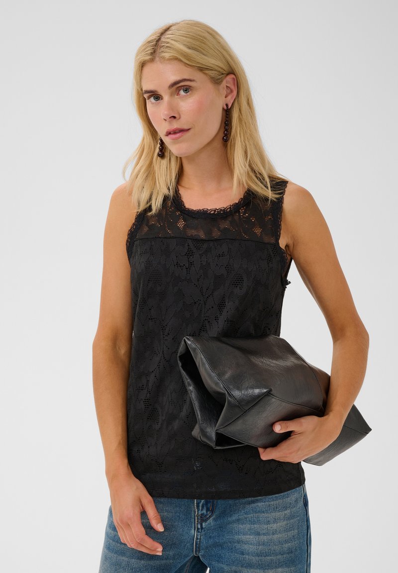 Femme blonde portant un haut en dentelle noire sans manches et un jean bleu tient une grande pochette en cuir noir pliée.
