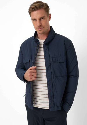 Mand iført navy quiltet jakke over hvid skjorte med tynde brune striber, stående mod en ensfarvet hvid baggrund.