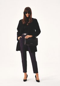 Cappotto nero sopra una maglietta nera, abbinato a pantaloni viola scuro con strisce laterali. Accessoriato con una borsa a tracolla nera e tacchi neri.