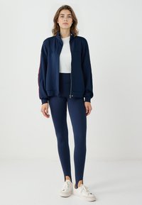 Giacca zip atletica navy con colletto alto, abbinata a leggings coordinati e sneakers bianche. Tessuto morbido, design aderente e tagli laterali.