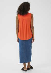 Top naranja sin mangas con un diseño de espalda fruncida, combinado con una falda larga de mezclilla azul y sandalias negras de plásticos. Textura suave, estilo casual.