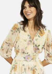 Õieprintiga maksikleit pehmes kollases toonis, millel on V-kaelus, rüüsärgid ja kuldsed detailid. Materjal on kerge ja läbipaistev.