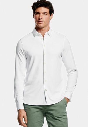 Chemise blanche à manches longues avec un col boutonné, en tissu lisse. Elle présente un ourlet droit et des boutons beige clair. Associée à un pantalon vert.