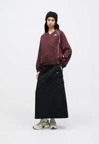 Vínový pulóver s dlhými rukávmi a bielym lemovaním, kombinovaný s čiernou sukňou dĺžky po kolená s vreckami. Oblečený so sivými teniskami a čiernou čiapkou.