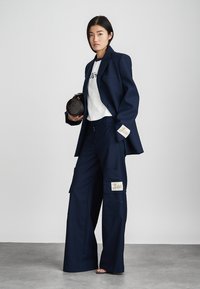 Blazer navy abbinato a una t-shirt grafica bianca e pantaloni cargo a gamba larga. I dettagli includono toppe sui pantaloni e una borsa nera rotonda.