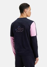 Sweat-shirt bleu marine avec des manches roses, présentant un logo texturé "Eden Park" au dos. Fabriqué dans un matériau doux avec une coupe décontractée.
