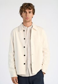 Krämfärgad jacka med krage, svarta knappar och bröstficka, lager-på-lager över en ljusrandig skjorta, bärs med mörka byxor.