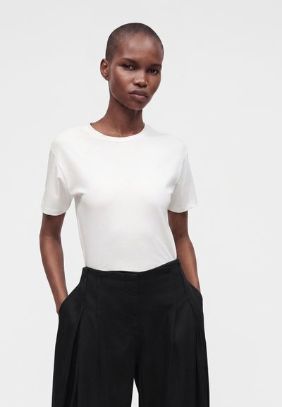 Femme aux cheveux courts portant un t-shirt blanc uni rentré dans un pantalon noir taille haute à larges jambes, les mains dans les poches.
