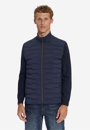 Jonge man met een marineblauw doorgestikt vest met rits over een overhemd met lange mouwen en blauwe spijkerbroek, staand tegen een effen achtergrond.