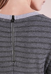 Dettaglio della parte superiore della schiena di una persona che mostra una cerniera nera su un maglione in maglia grigio e nero con colletto grigio chiaro.