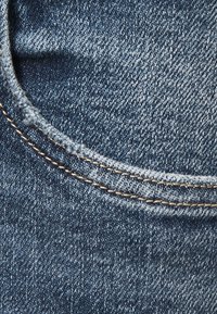 Primer plano de tela de mezclilla azul que muestra el borde curvado del bolsillo con costura beige y textura de tejido visible.