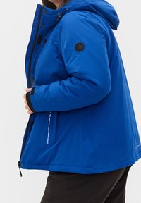 Chaqueta azul aislante con capucha, con detalles en negro y un parche de logo. Costura blanca a lo largo del lateral. Textura suave y material duradero.