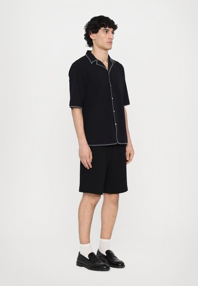Short-sleeve button-up shirt in black fabric with contrasting white stitching, paired with black shorts and black slip-on shoes.