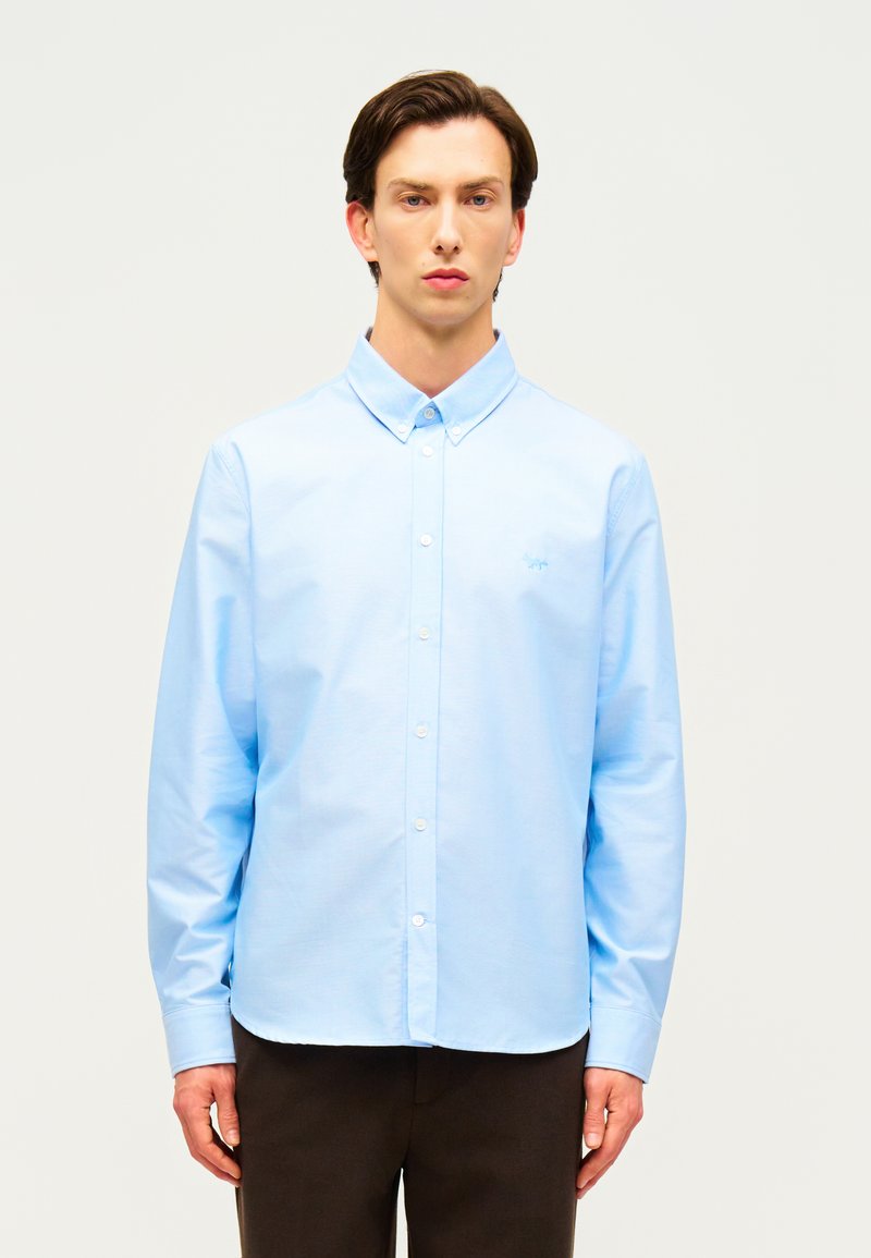 Νεαρός άνδρας που φοράει ανοιχτό γαλάζιο πουκάμισο με κουμπιά και σκούρο παντελόνι, στέκεται μπροστά σε απλό ανοιχτό φόντο, κοιτάζοντας προς τα εμπρός.