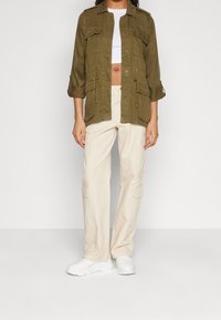 Oliwkowa kurtka użytkowa z przednimi kieszeniami, zestawiona z beżowymi spodniami cargo i białymi trampkami, prezentująca luźny krój i lekką tkaninę.