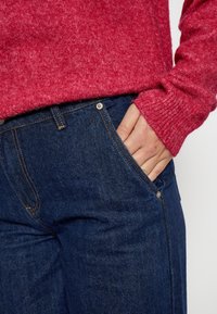 Crvena teksturirana pletena dolčevita s dugim rukavima preko tamnoplavih traperica. Traperice imaju prednje džepove i kontrastne narančaste šavove.