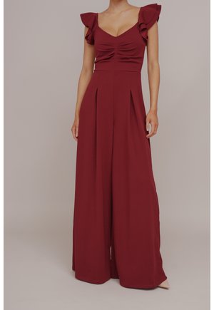 Kobieta ubrana w bezrękawnikowy bordowy kombinezon z falbanowymi ramiączkami i spodniami o szerokich nogawkach, stojąca na jednolitym tle.