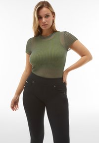 Vihreä raidallinen lyhythihainen paita, valmistettu läpikuultavasta materiaalista, yhdistetty mustiin istuviin housuihin, joissa on taskudetaljit ja sileä vyötärönauha.