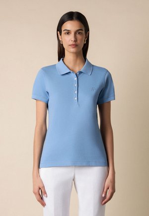 Vrouw draagt een lichtblauw poloshirt met korte mouwen en witte broek, staand tegen een effen beige achtergrond.