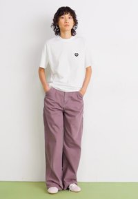 Giovane con capelli ricci che indossa una t-shirt bianca con logo a cuore, pantaloni larghi mauve e sneakers rosa chiaro, in piedi con le mani nelle tasche.