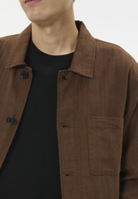 Muškarac u smešnom jakni sa dugmi smeđe boje preko crne majice s okruglim izrezom, stoji naslonjen na jednostavnu svijetlu pozadinu.