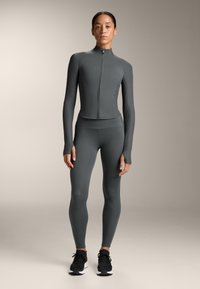 Träningsset i mörkgrått, bestående av en tight jacka med hög krage och hel dragkedja, tillsammans med matchande leggings och svarta sneakers.