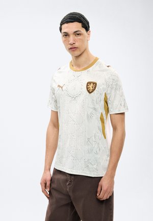 Hombre joven con cabello corto y oscuro, vistiendo una camiseta deportiva blanca con textura y detalles dorados, y pantalones oscuros, de pie frente a un fondo liso.