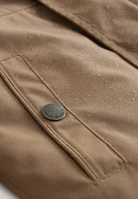 Brown waterproof fabric featuring a front pocket and a metallic button. Surface displays droplets, indicating moisture-resistance.