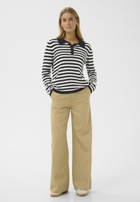 Donna in piedi con le mani nelle tasche, che indossa una maglietta a maniche lunghe bianco e nero a righe e pantaloni larghi beige, completi di scarpe bianche.